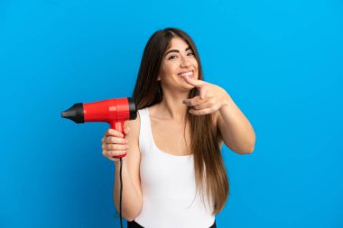 Mavi arka planda saç kurutma makinesi tutan beyaz bir kadın mutlu bir ifadeyle öne bakıyor.
