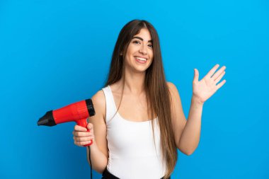 Beyaz bir kadın elinde saç kurutma makinesiyle mavi arka planda izole edilmiş bir şekilde elinde mutlu bir ifadeyle selam veriyor.