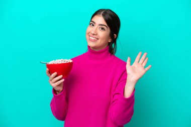 Beyaz bir kadın elinde bir kase mısır gevreği tutarken mavi arka planda mutlu bir ifadeyle selam veriyor.