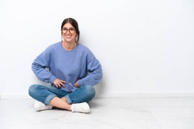 Beyaz arka planda izole bir şekilde yerde oturan genç beyaz kadın kolları kalçasında ve gülümsüyor.