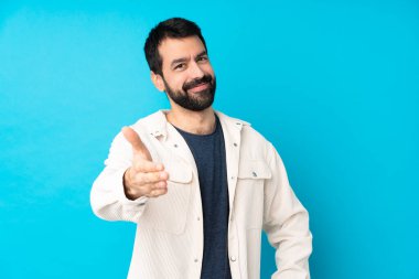 Beyaz kadife ceketli yakışıklı genç adam izole edilmiş mavi arka planda iyi bir anlaşma için el sıkışıyor.