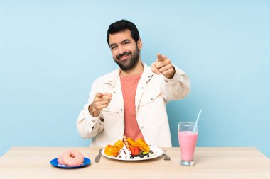 Kahvaltıda waffle yiyen ve milkshake içen bir adam gülümserken parmağıyla seni işaret ediyor.