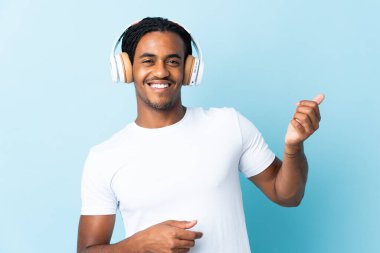 Mavi arka planda izole edilmiş örgülü genç Afrikalı Amerikalı adam müzik dinliyor ve gitar hareketi yapıyor.