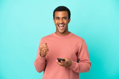 İzole edilmiş mavi arka planda, telefonu zafer pozisyonunda olan Afro-Amerikalı yakışıklı adam.