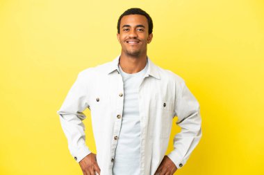 Sarı arka planda, kolları kalçasında gülümseyerek poz veren Afro-Amerikalı yakışıklı bir adam.