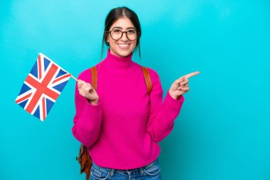 İngiliz bayrağını elinde tutan beyaz genç bir bayan mavi arka planda izole edilmiş olarak parmak kaldırıyor.