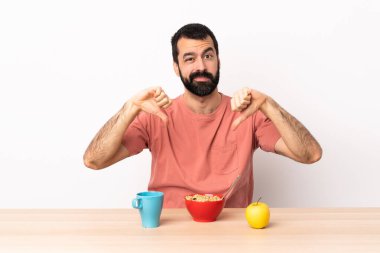 Masada kahvaltı yapan beyaz bir adam baş parmağını iki eliyle aşağı gösteriyor..