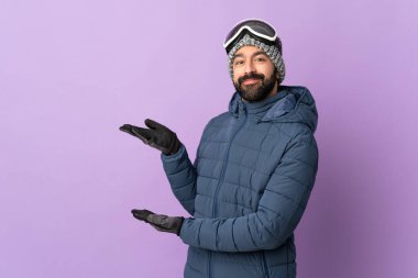 İzole edilmiş mor arka planda snowboard gözlüklü kayakçı. Gelmesi için ellerini yana uzatıyor.