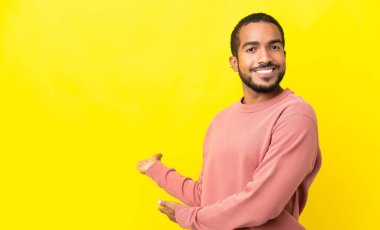 Sarı arka planda izole edilmiş genç latin adam, davet edilmek için ellerini yana uzatıyor.