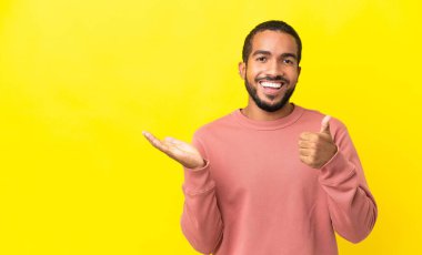 Sarı arka planda izole edilmiş genç bir latin adam. Avucunun içinde hayali bir şey tutuyor. Bir ilan vermek için.