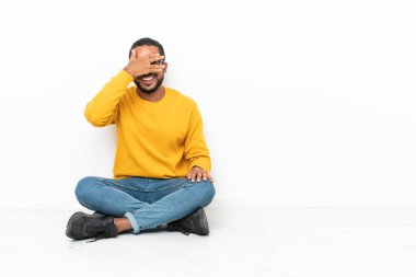 Genç Ekvadorlu adam, beyaz duvarda izole bir şekilde yerde oturuyor gözleri kaplıyor ve gülümsüyor.