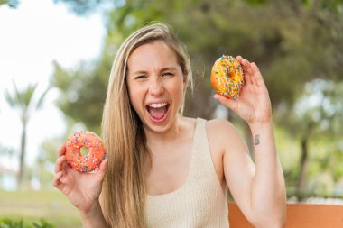 Açık havada genç sarışın bir kadın elinde mutlu bir ifadeyle donut tutuyor.