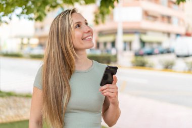 Açık havada cep telefonu kullanan genç sarışın kadın gülümserken yukarı bakıyor.