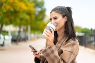 Dışarıda cep telefonu kullanan ve elinde kahve tutan genç ve güzel bir kadın.