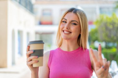 Açık havada gülümseyen ve zafer işareti gösteren güzel sarışın bir kadın.
