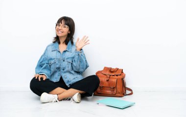 Genç bir kız öğrenci, beyaz arka planda tek başına oturmuş mutlu bir ifadeyle selamlıyor.