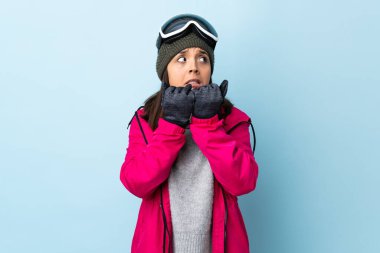 Snowboard gözlüklü karışık kayakçı kız izole edilmiş mavi arka planda gergin ve elleri ağıza vermeye korkuyor..