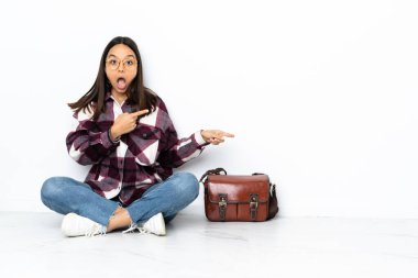 Genç bir bayan öğrenci yerde oturuyor, şaşırmış ve işaret ediyor.