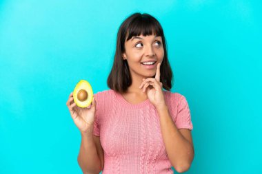 Mavi arka planda avakado tutan genç melez kadın yukarı bakarken bir fikir düşünüyor.