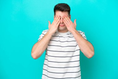 Genç beyaz adam mavi arka planda izole edildi. Gözlerini elleriyle kapadı.