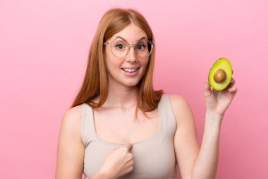 Elinde avakado tutan kızıl saçlı genç bir kadın. Pembe arka planda, yüzünde sürpriz bir ifade var.
