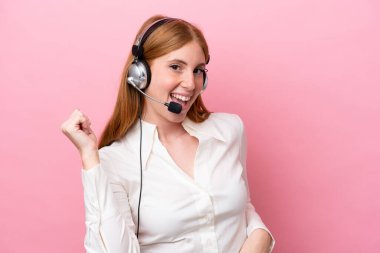 Tele pazarlamacı kızıl saçlı kadın pembe arka planda izole edilmiş bir kulaklıkla gitar jesti yapıyor.