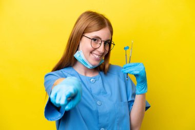 Genç kızıl saçlı dişçi kadın, sarı arka planda izole edilmiş mutlu bir ifadeyle ön tarafı işaret ediyor.