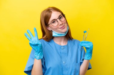 Genç kızıl saçlı dişçi kadın sarı arka planda izole edilmiş mutlu ve parmaklarıyla üçe kadar sayıyor