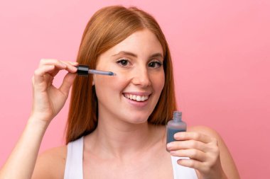 Genç kızıl saçlı kadın pembe arka planda izole edilmiş, elinde bir serum tutarken gülümsüyor. Portreyi kapat