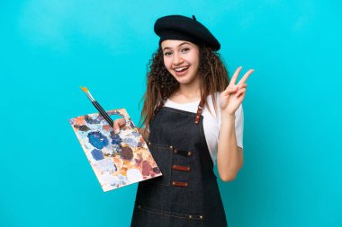 Genç sanatçı Arap kadın mavi arka planda izole edilmiş bir palet tutuyor gülümsüyor ve zafer işareti gösteriyor.