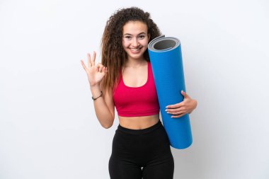 Genç Arap spor kadını yoga derslerine giderken beyaz arka planda izole edilmiş bir minder tutuyor ve parmaklarıyla işaret veriyor.