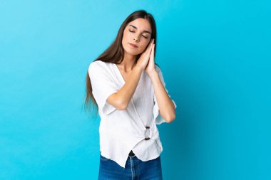 Mavi arka planda izole edilmiş genç beyaz kadın uyku jesti yapıyor.