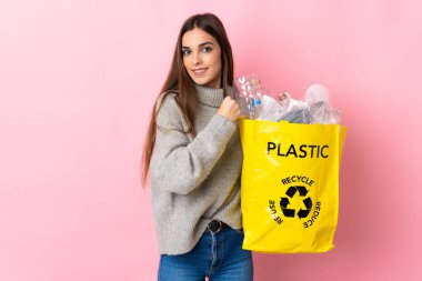 Pembe arka planda izole edilmiş bir şekilde zaferi kutlamak için elinde plastik şişelerle dolu bir çanta tutan beyaz bir kadın.