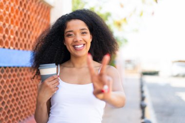 Genç Afrikalı Amerikalı bir kadın açık havada kahve tutuyor gülümsüyor ve zafer işareti gösteriyor.