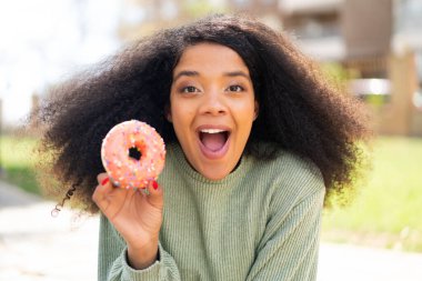 Genç Afro-Amerikalı bir kadın elinde sürpriz ve şok edici bir yüz ifadesiyle açık havada donut tutuyor.