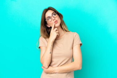 Kafkasyalı genç bir kadın mavi arka planda izole edilmiş, yukarı bakarken şüpheleri var.