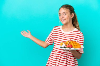 Mavi arka planda izole edilmiş waffle 'ları tutan küçük beyaz kız davet etmek için ellerini yana uzatıyor.