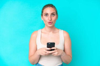 Mavi arka planda izole edilmiş genç beyaz kadın kameraya bakarken şaşırmış bir ifadeyle cep telefonunu kullanıyor.
