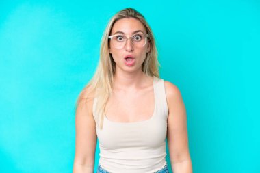 Mavi arka planda gözlüklü ve şaşkın bakışlı genç beyaz kadın.