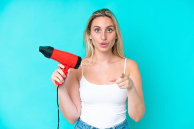 Mavi arka planda, elinde saç kurutma makinesi tutan beyaz bir kadın şaşırmış ve ön tarafı işaret ediyor.