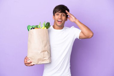 Elinde alışveriş poşeti tutan genç bir adam. Mor arka planda, sürpriz bir ifadeyle izole edilmiş.