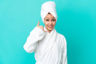 İzole edilmiş mavi arka planda bornozlu genç sarışın kız telefon jesti yapıyor. Bana geri dönüş işareti ver.