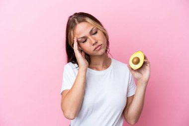 Avokado tutan genç Rus kadın pembe arka planda baş ağrısıyla izole edilmiş.