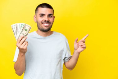 Young caucasian man taking a lot of money isolated on yellow background pointing finger to the side