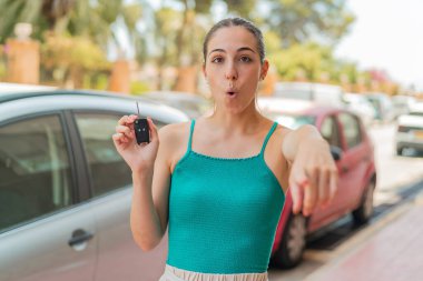 Dışarıda arabanın anahtarlarını tutan genç ve güzel bir kadın şaşırmış ve ön tarafı işaret ediyor.
