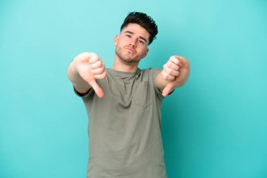 Mavi arka planda izole edilmiş genç beyaz bir adam baş parmağını iki eliyle aşağı gösteriyor.