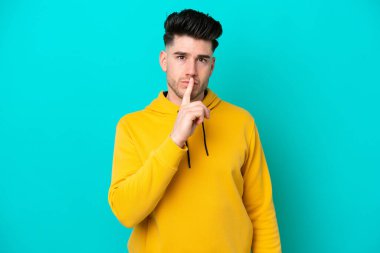 Mavi arka planda izole edilmiş genç beyaz adam sessiz bir jest işareti gösteriyor. Parmağını ağzına sokuyor.