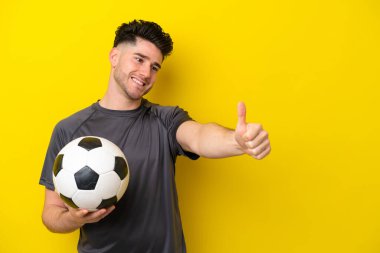 Yakışıklı genç futbolcu sarı arka planda izole edilmiş bir şekilde baş parmak hareketi yapıyor.