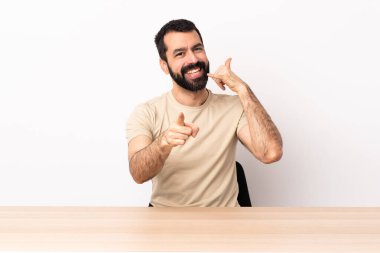 Masada sakallı, beyaz bir adam telefon jesti yapıyor ve ön tarafı işaret ediyor..