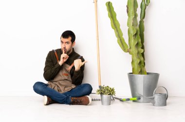 Bahçıvan içeride yerde oturuyor, yan tarafı işaret ediyor ve sessiz jest yapıyor.